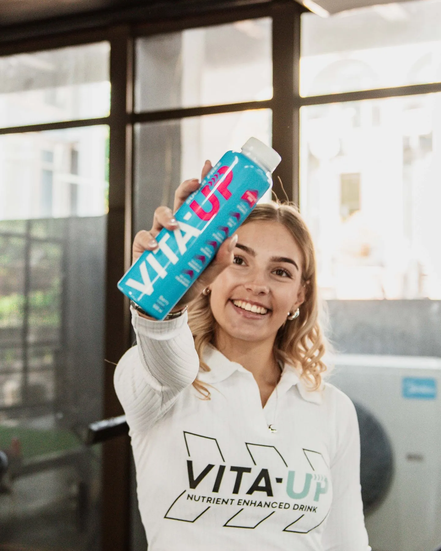 Smiling woman in a Vita-Up top holding up a Berry bottle in the gym